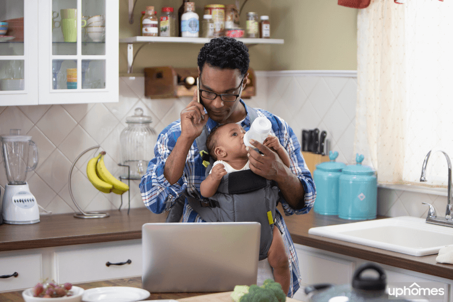 Hardworking single Dad feeding baby in a baby carrier while on the phone, searching the computer and multitasking as a single parent with a ton of things going on
