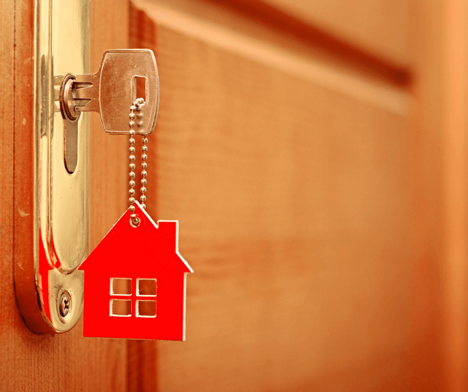 Extreme close-up of a key inserted into the lock of a house’s front door; the keychain, hanging below the key, is a red cutout in the shape of a house.